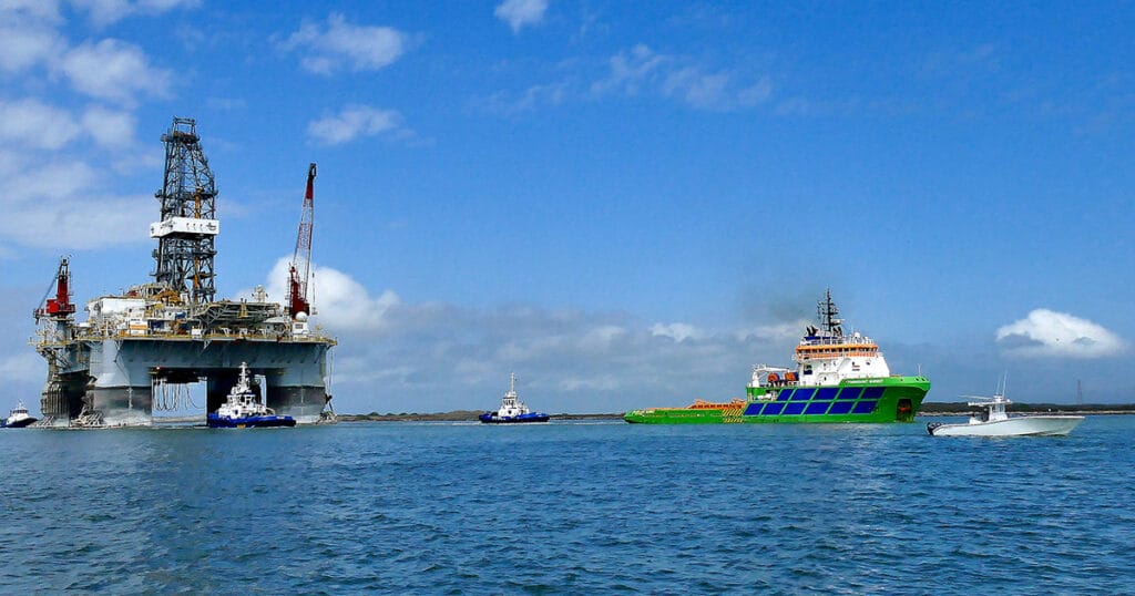 Oil Drilling platform being towed in channel to Gulf of America by multiple boats.
