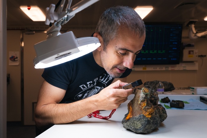 Chief Scientist Aaron Micallef (Monterey Bay Aquarium Research Institute) studies a rock from a massive sulfide deposit. While hydrothermal vents are individual features, with brief lifespans of thousands of years, these massive sulfide deposits are formed on a much larger scale and over far greater timescales: they are highly significant, rare to find, and of great interest to the science team. (Image credit: Alex Ingle, Schmidt Ocean Institute)