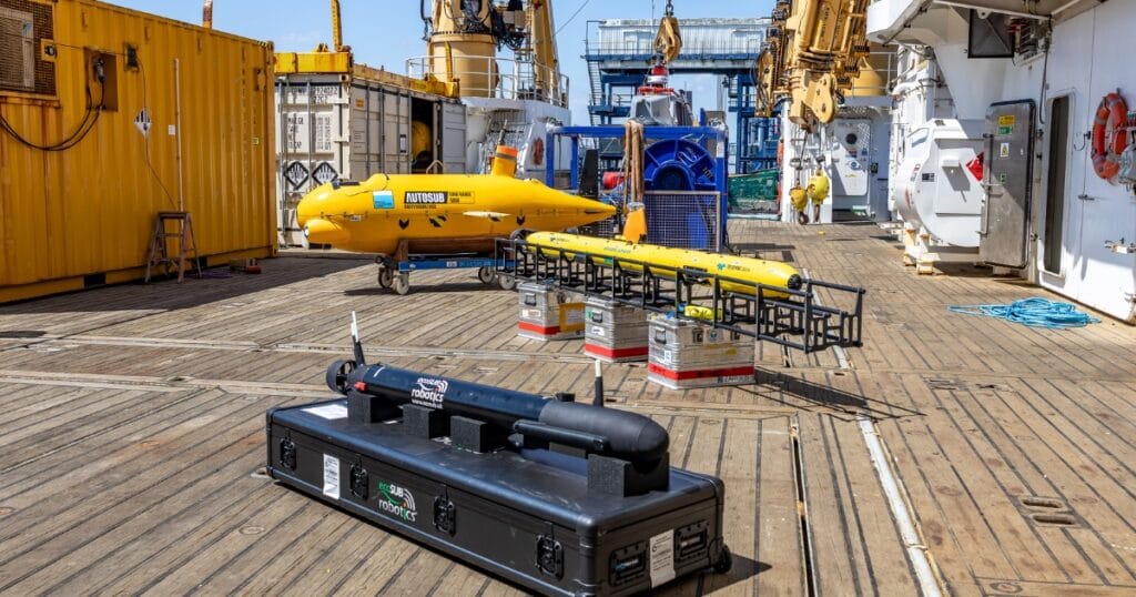 Three autonomous underwater vehicles (AUVs) of varying sizes and colors, along with shipping containers and cranes, on the wooden deck of a ship under a clear blue sky.