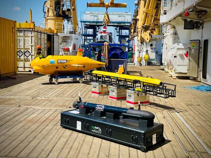 Three autonomous underwater vehicles (AUVs) – a large yellow AUTOSUB, a long yellow Teledyne Gavia, and a black ecoSUB Robotics vehicle – displayed on the wooden deck of a ship with cranes and containers in the background.